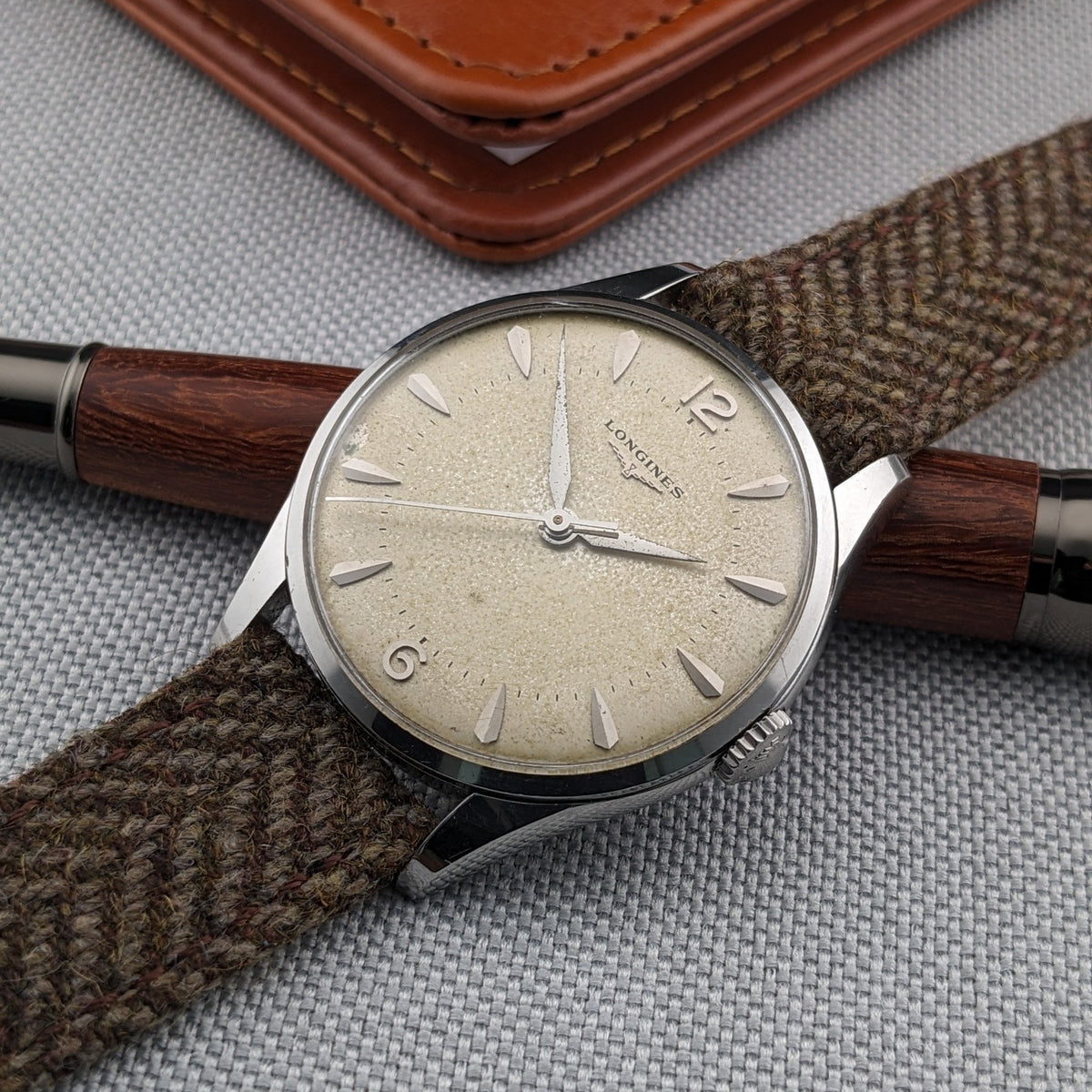 Longines Classic reference 8135-5 rests across a dark wood pen on gray fabric, with its cream dial and brown woven strap centered.