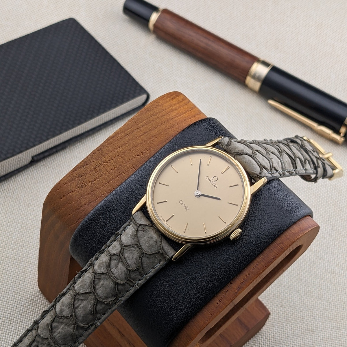 Omega De Ville reference 111 0156 displayed on a black-and-wood watch stand, with a dark notebook and pen behind it.