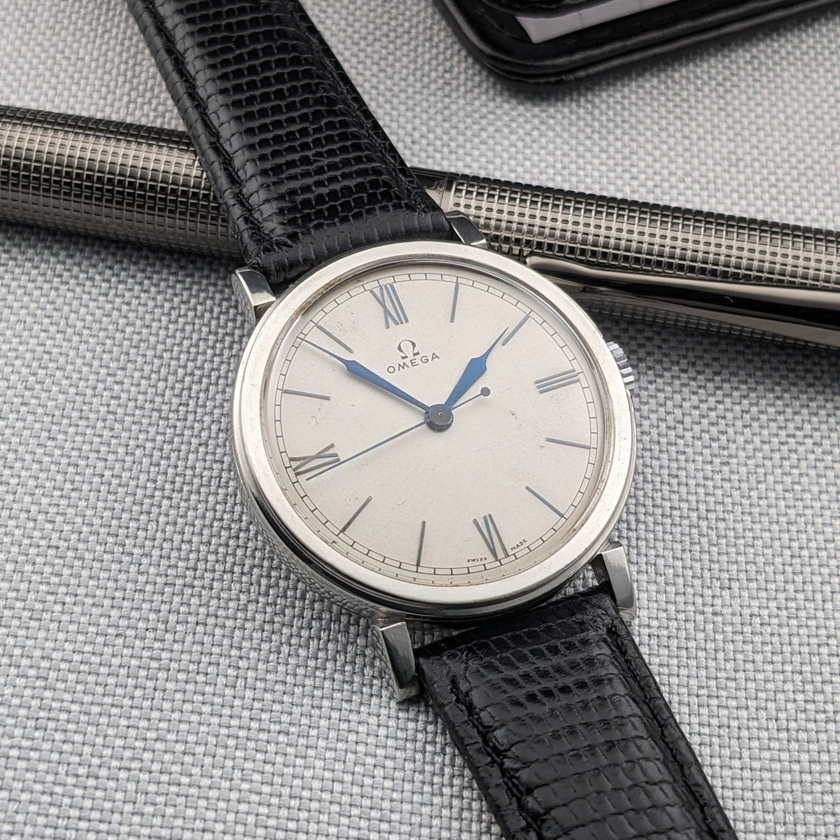 Angled Omega watch view showing the light dial, blue hands, Roman numerals and black strap over a silver pen.