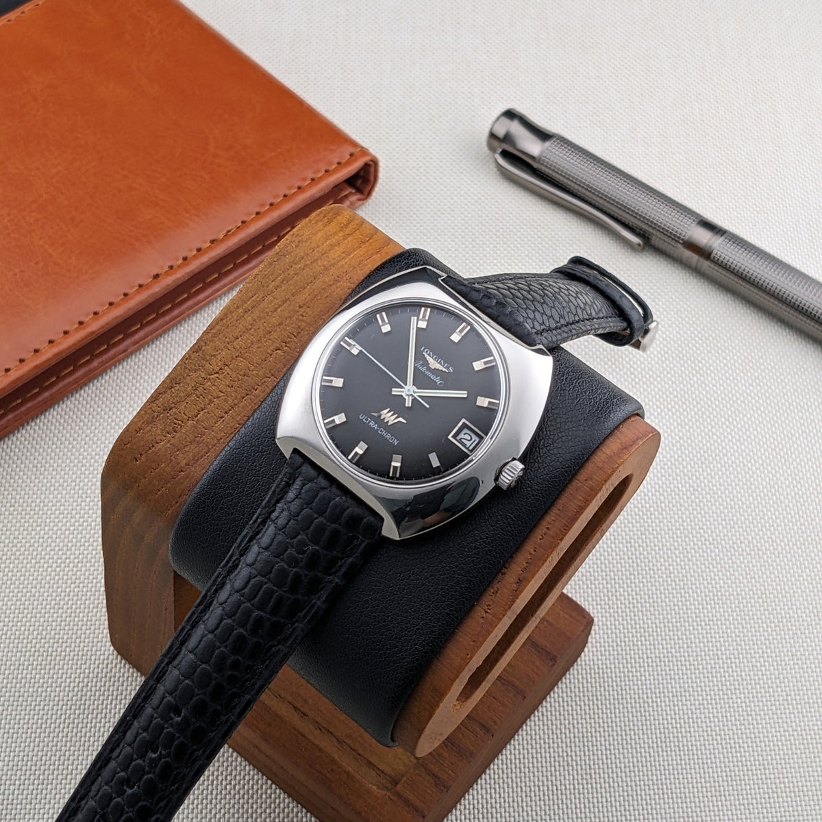 Longines Ultrachron reference 8429 resting on a wooden watch stand with black cushion, brown wallet and silver pen in the background.