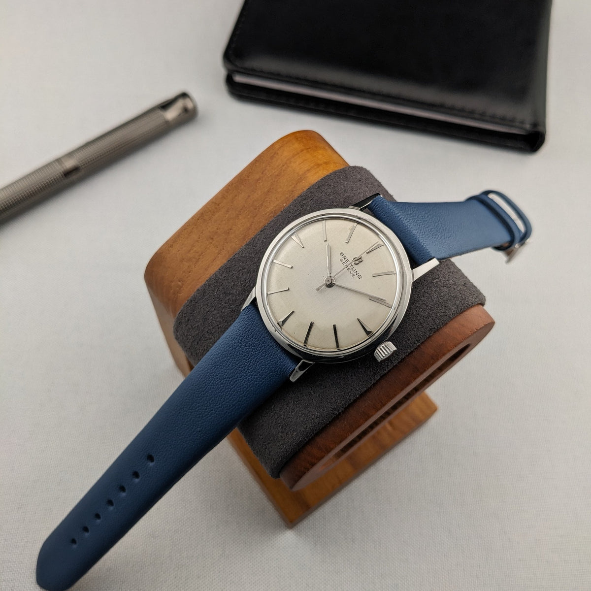 Breitling Automatic watch placed on a wooden watch stand, with its blue strap extended beside a black case and silver pen.
