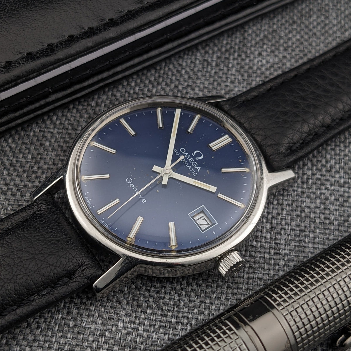 Angled Omega Geneve Automatic reference 166.0163 with blue dial, date window, applied markers and black leather strap on gray fabric.