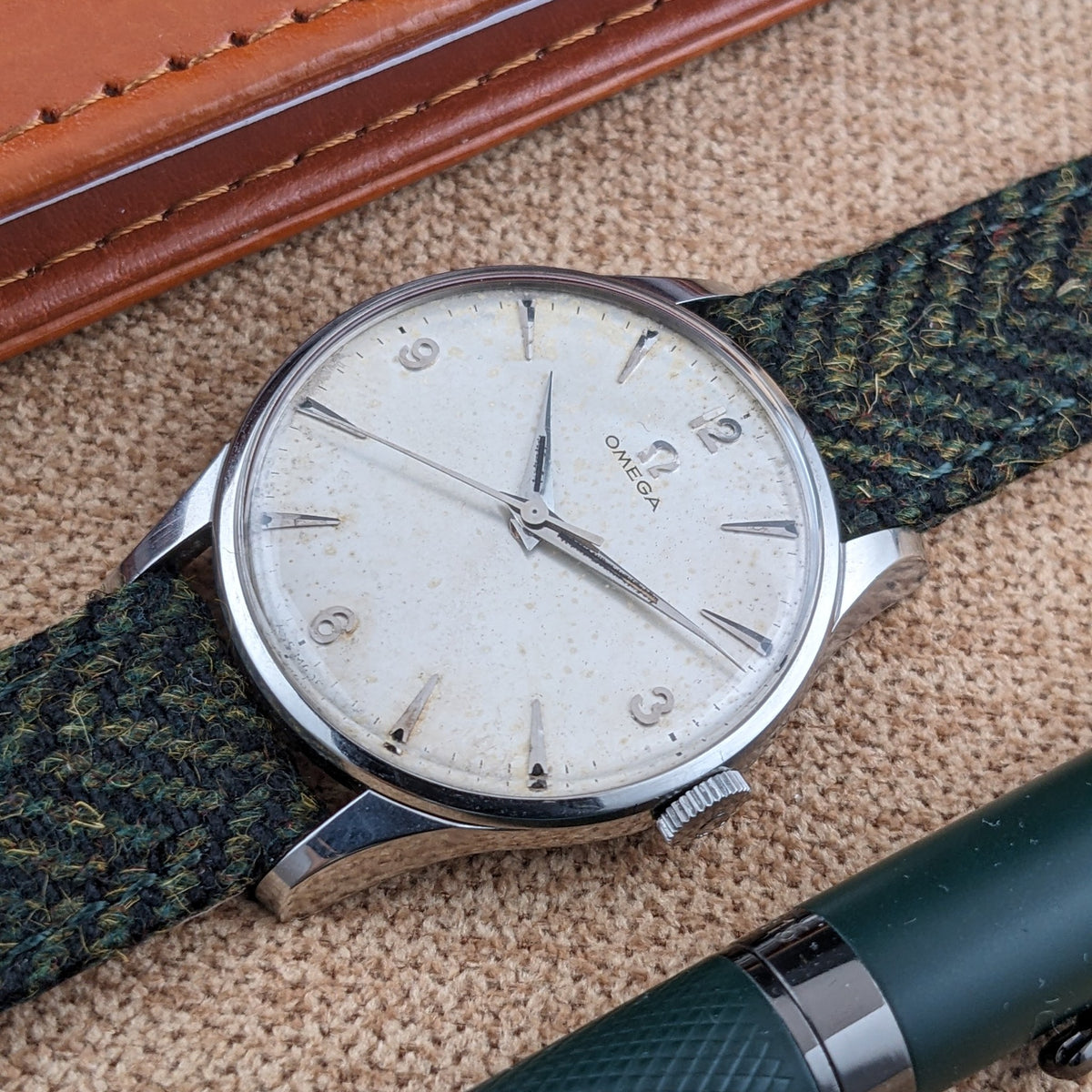 Omega Classic Referenz 2810-2 SC diagonal gezeigt mit hellem Zifferblatt mit Ziffern, grünem Armband, braunem Notizbuch und dunklem Stift.