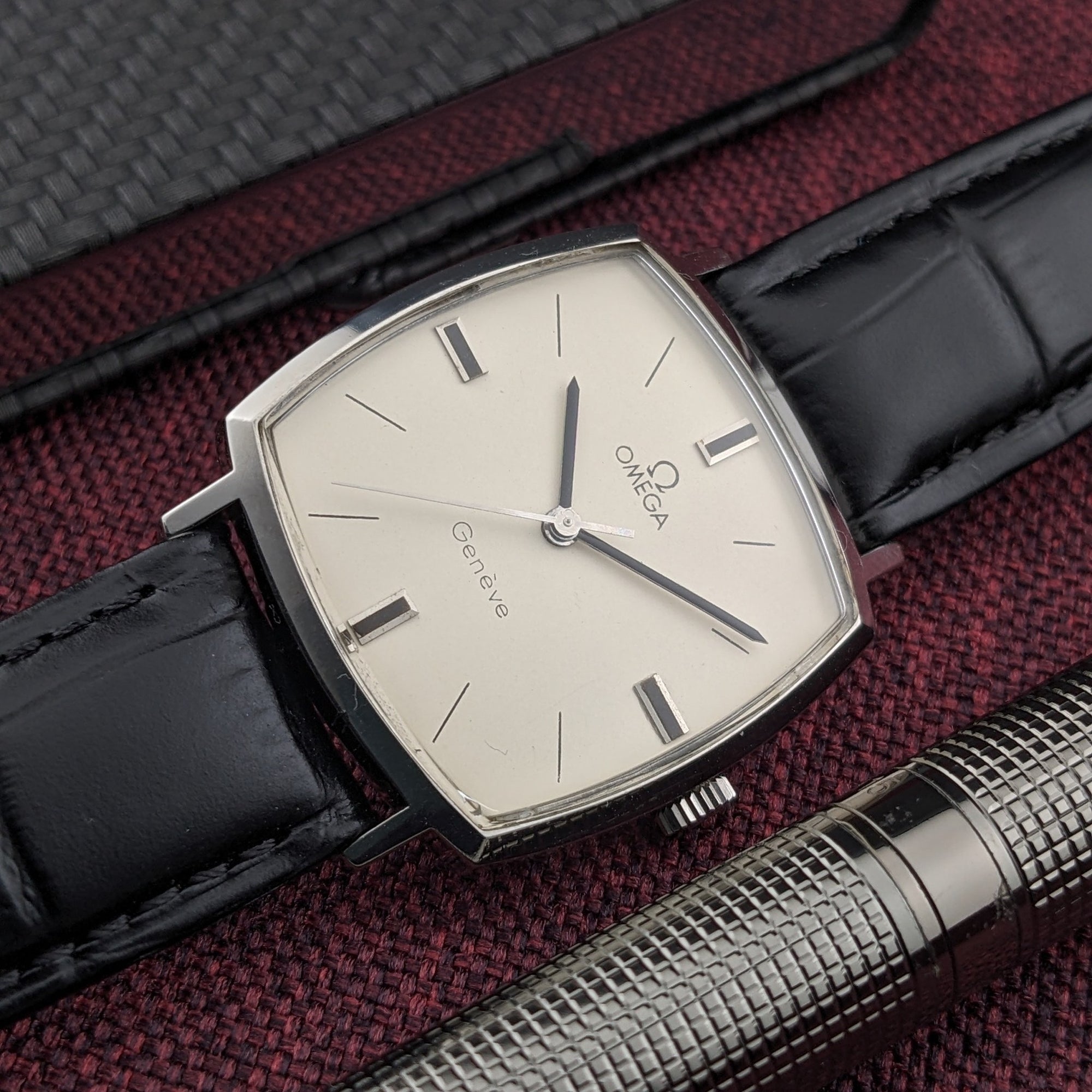 Omega Genève reference 131.022 displayed diagonally with its cream Genève dial, silver case, black strap, and a textured pen alongside.
