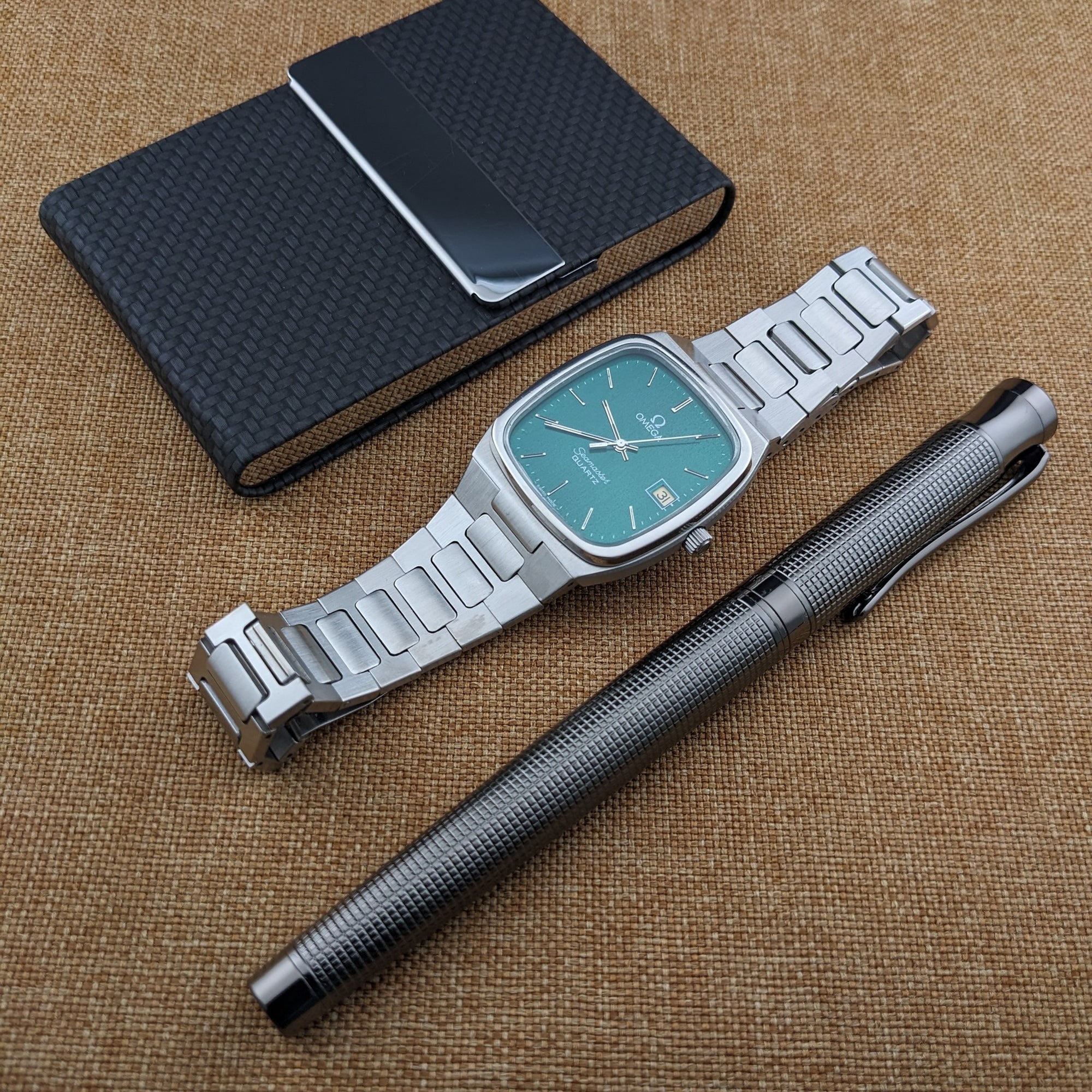 Omega Seamaster reference 196.0166 laid out with its open bracelet, black textured notebook, and black pen on tan fabric.