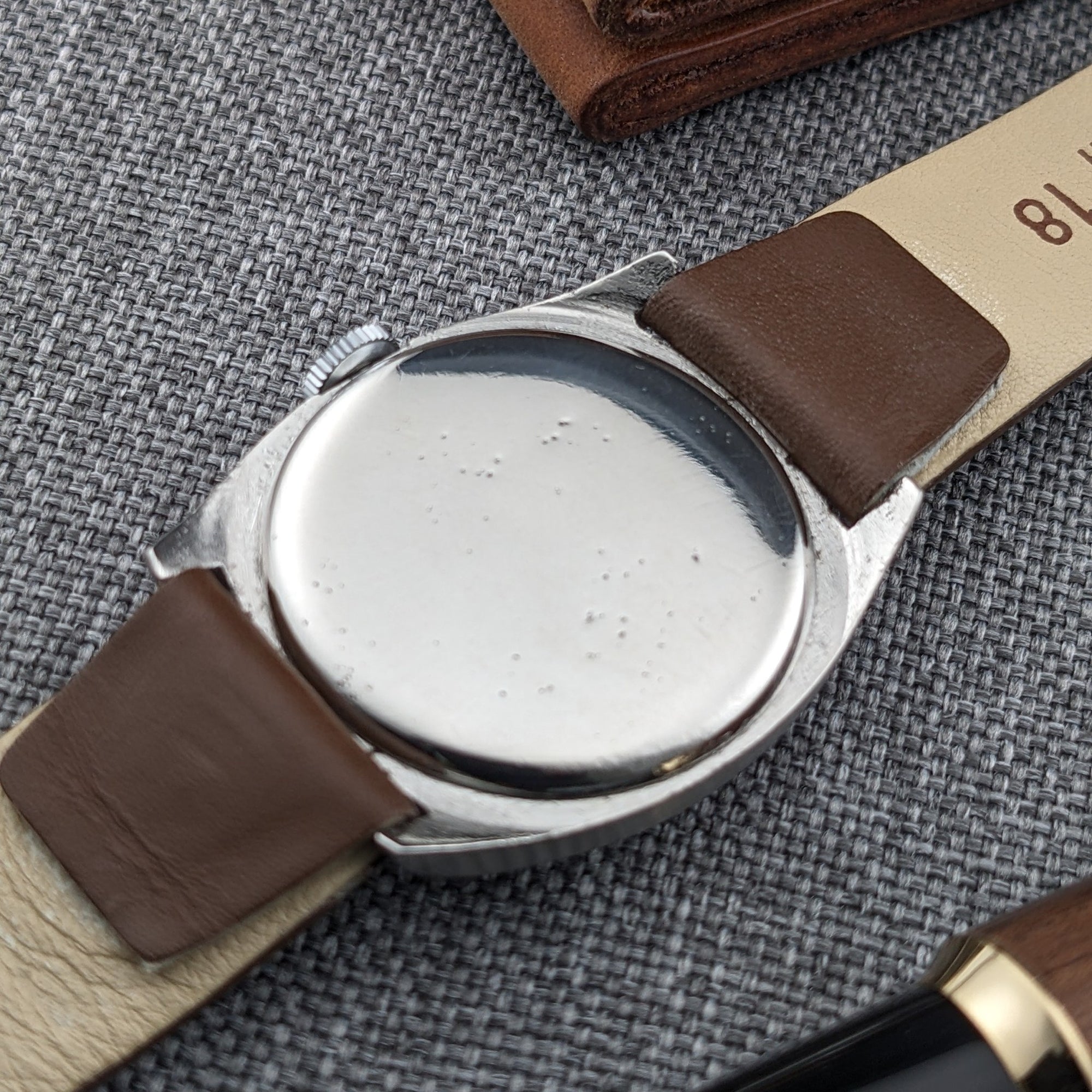 Rear view of an Omega watch showing a smooth round caseback, brown strap, and gray woven fabric background.