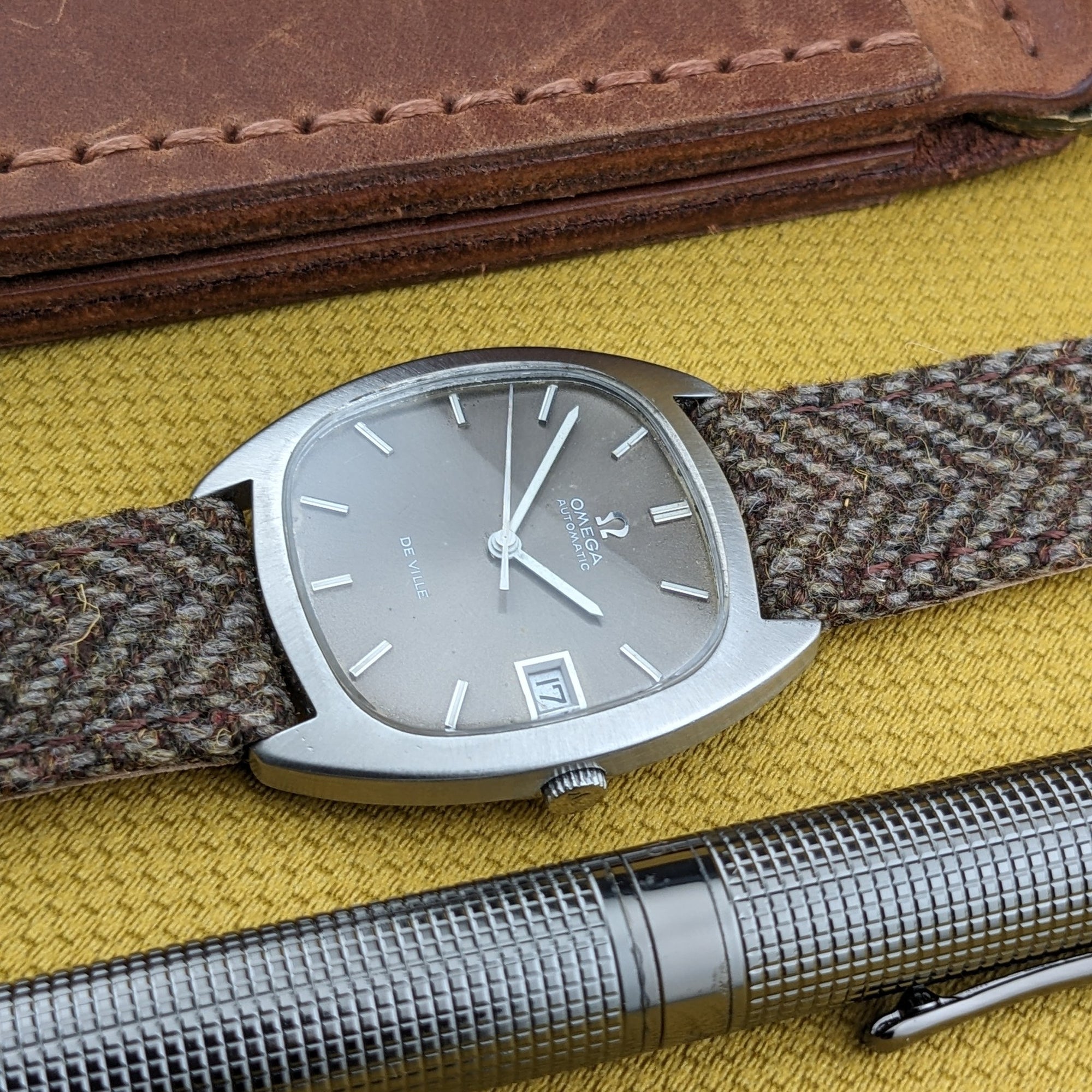 Omega Dr Ville Automatic ref. 162.045 lies beside a metal pen and leather wallet, with its silver dial and date window visible.