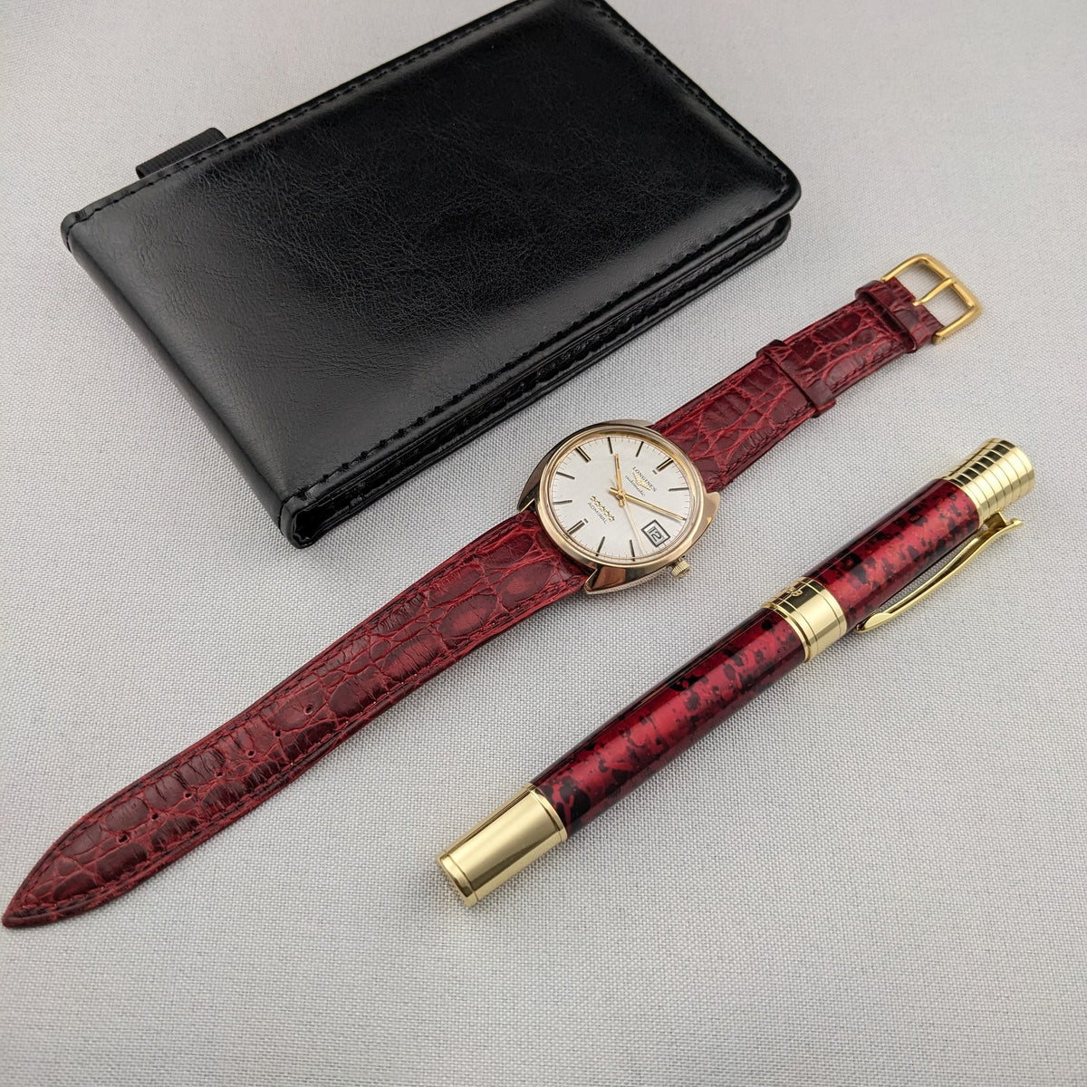 Longines Admiral Referenz 8343-1 neben einem schwarzen Etui und einem burgunder-goldenen Stift, mit dem vollständigen burgunderroten Armband sichtbar.