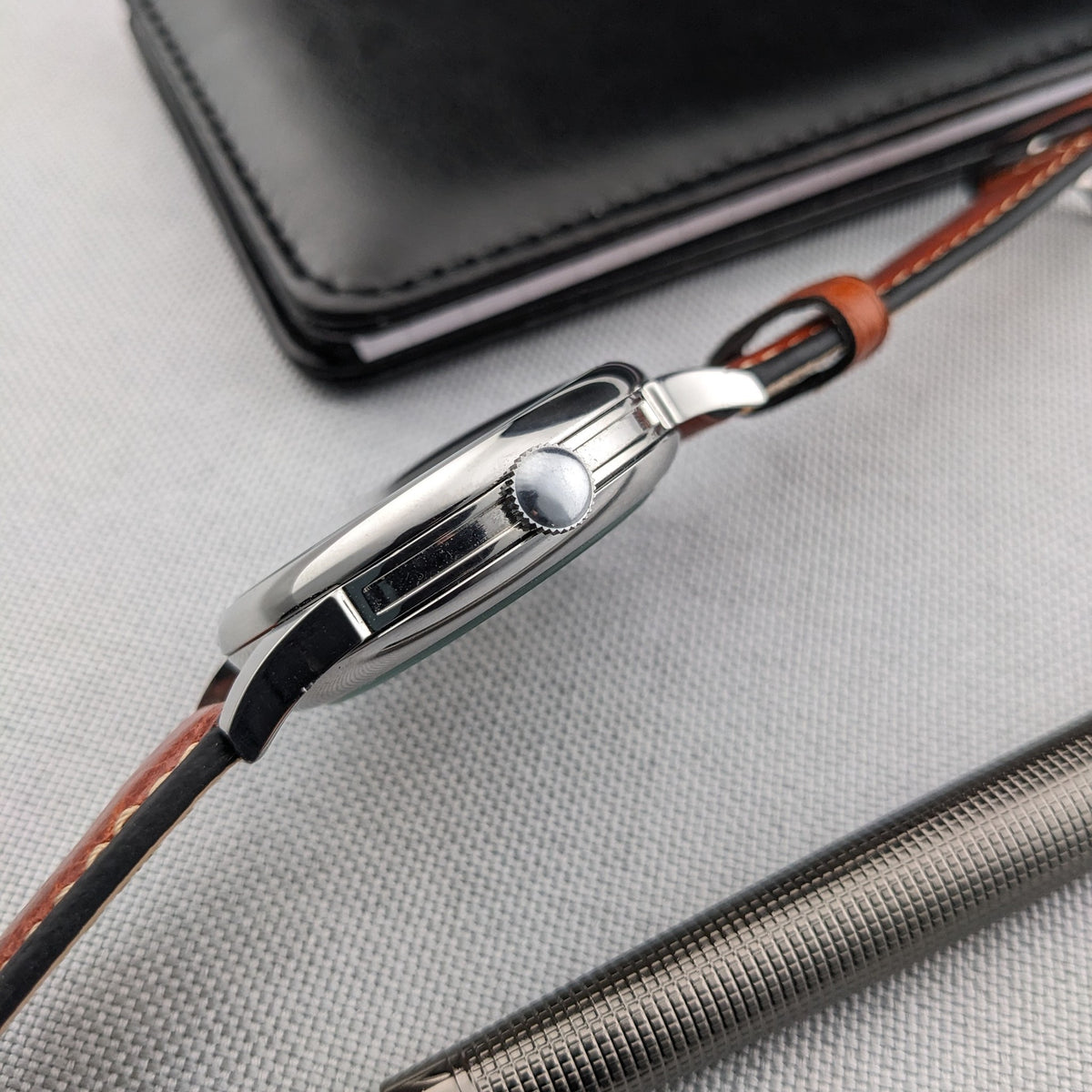 Seitenprofil der Omega-Armbanduhr mit poliertem Gehäuserand, Krone, Bandanstößen und braunem Lederarmband neben einem silbernen Stift.
