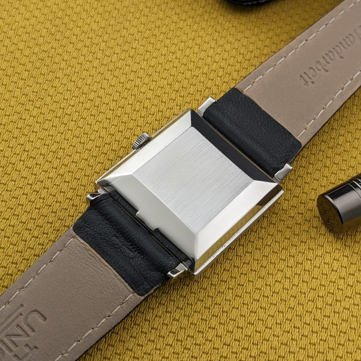 Rückansicht der Omega De Ville Automatik 161.014 mit gebürstetem rechteckigem Gehäuseboden, hellem Armbandunterseite und senffarbenem Hintergrund.
