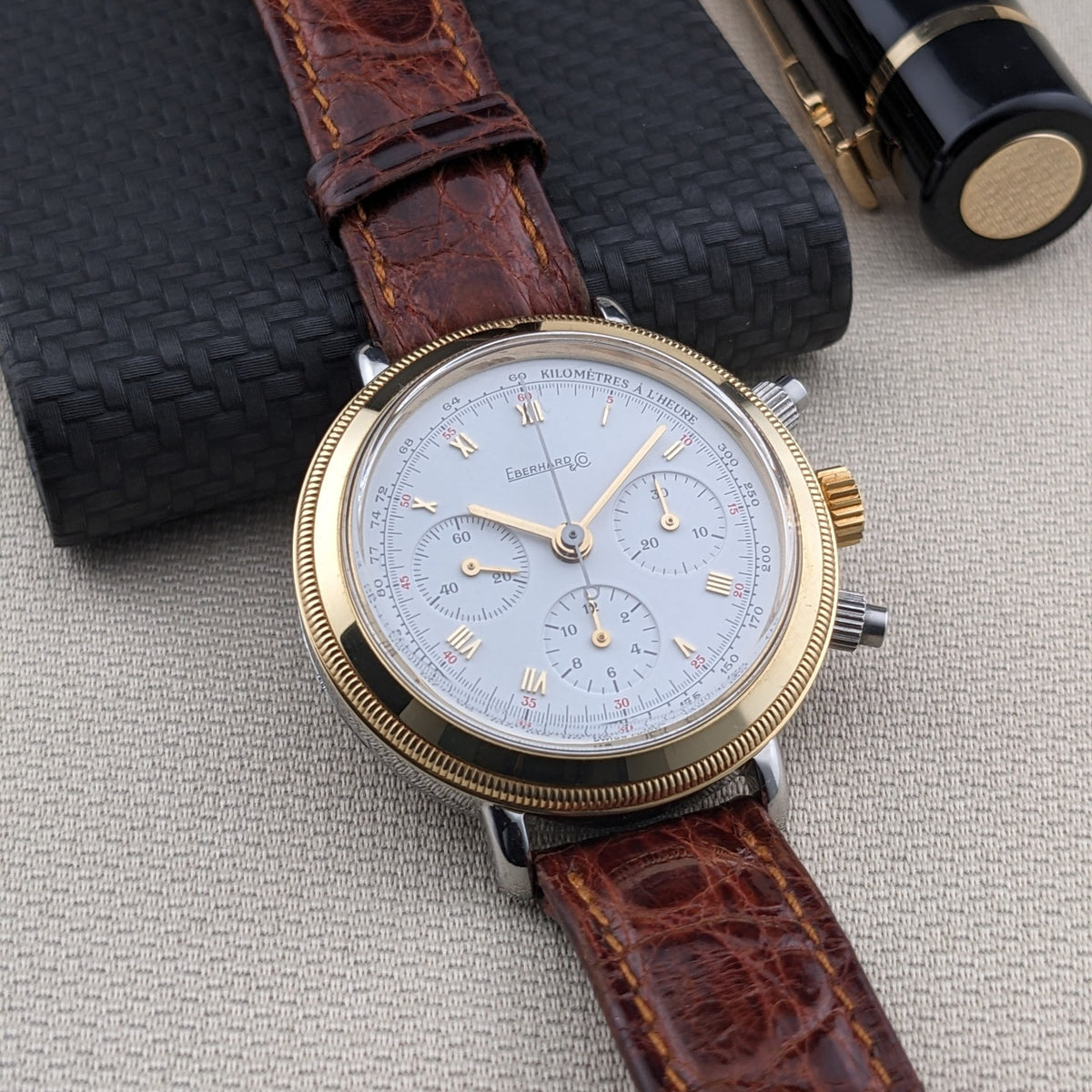 Eberhard & Co.. Godron Chrono reference 32031 lies on light fabric beside a black textured notebook and black fountain pen.