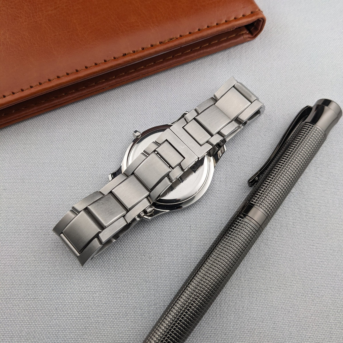 Montre Movado vue de l'arrière à côté d'un stylo texturé foncé et d'un étui en cuir brun, avec le bracelet en acier inoxydable ouvert.