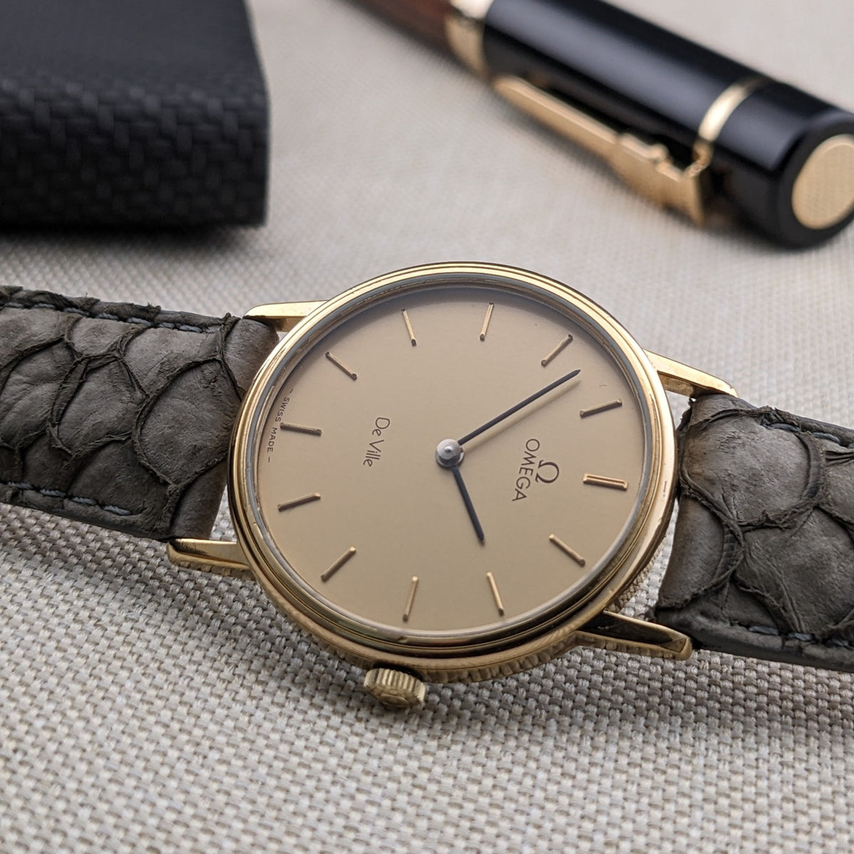 Close angled view of the Omega De Ville reference 111 0156 cream dial, gold-tone case, blue hands, crown, and gray strap.