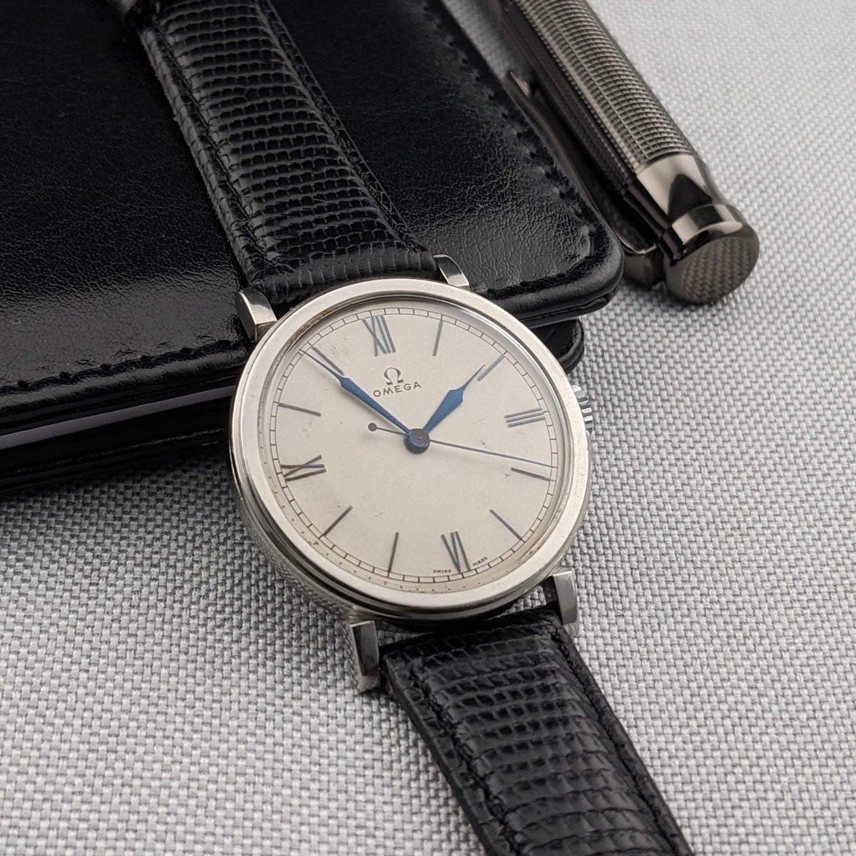 Omega watch positioned over a black wallet with a silver pen nearby, showing the dial and black textured strap.