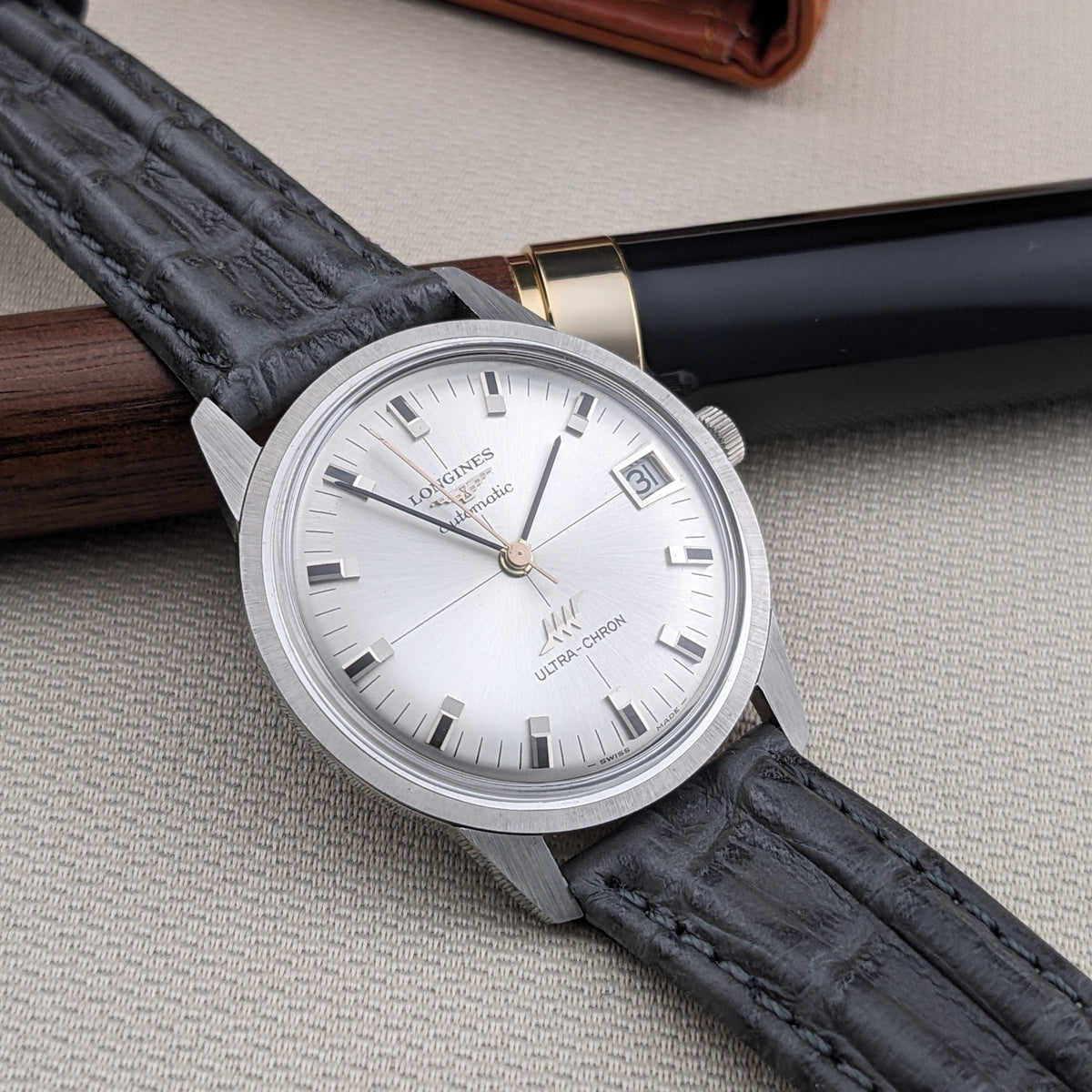 Longines Ultra-Chron 7827 angled across a wooden pen, showing the silver dial, date aperture, crown, and textured black strap.
