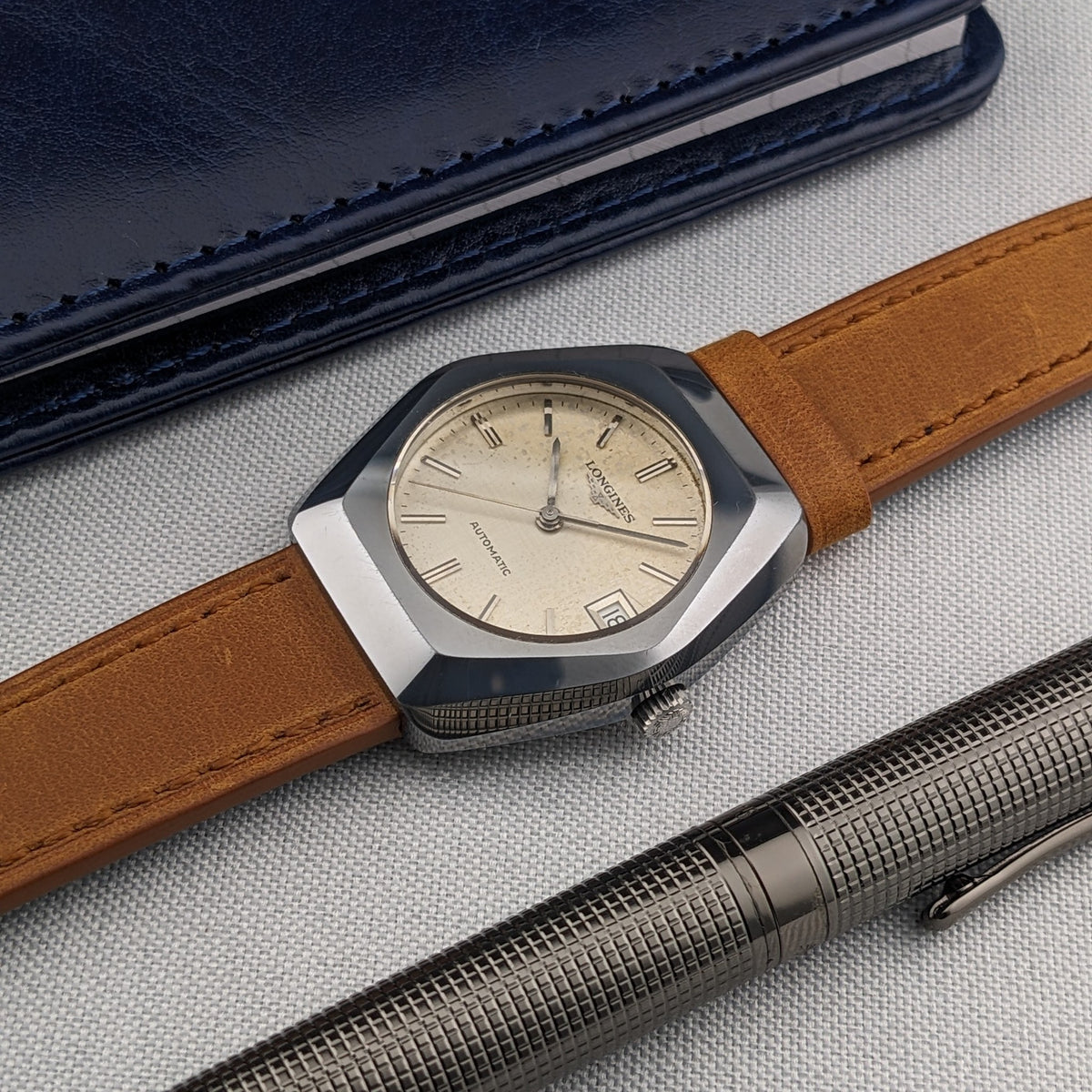LONGINES Automatic reference 3042-633 shown diagonally with cream dial, brown strap, blue case and textured metal pen.