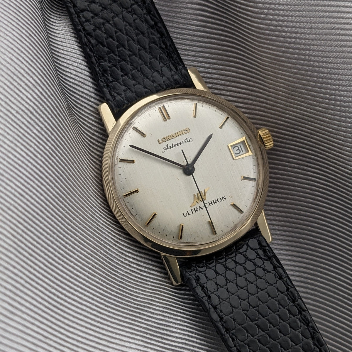 Longines Ultra-Chron 69-556AC resting on ribbed gray fabric, with cream dial, gold-tone markers, date window, and black textured strap.