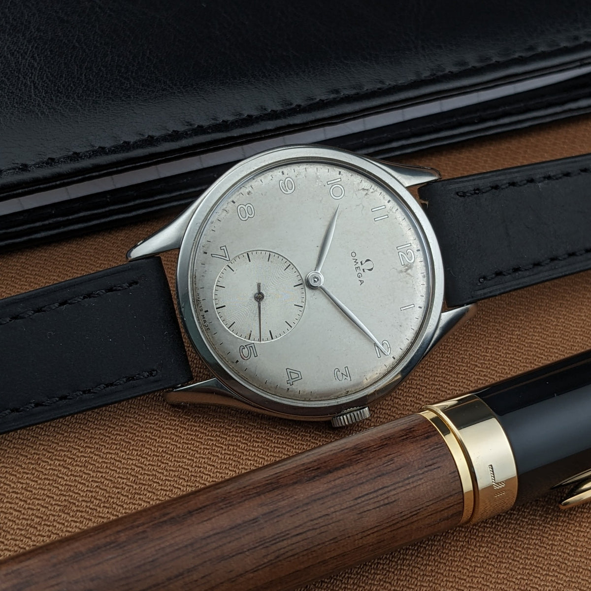 Omega Jumbo reference 2505-5 lying beside a black wallet and wooden pen, with its Arabic-numeral dial and small-seconds display visible.