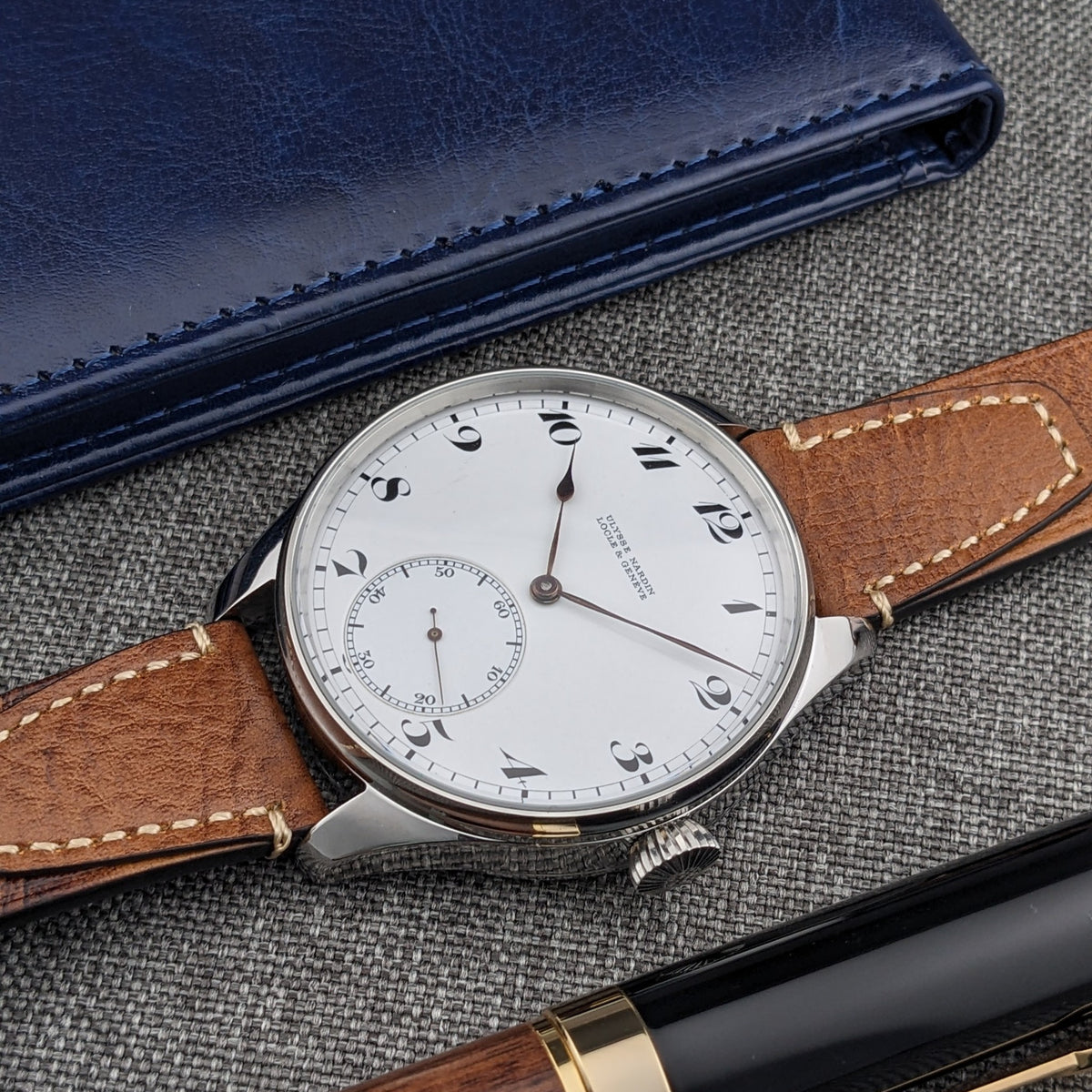 Ulysse Nardin कलाई घड़ी तिरछी दिखाई गई, जिसमें सफेद डायल और भूरा पट्टा, नीले केस और काले लेखन उपकरण के साथ।