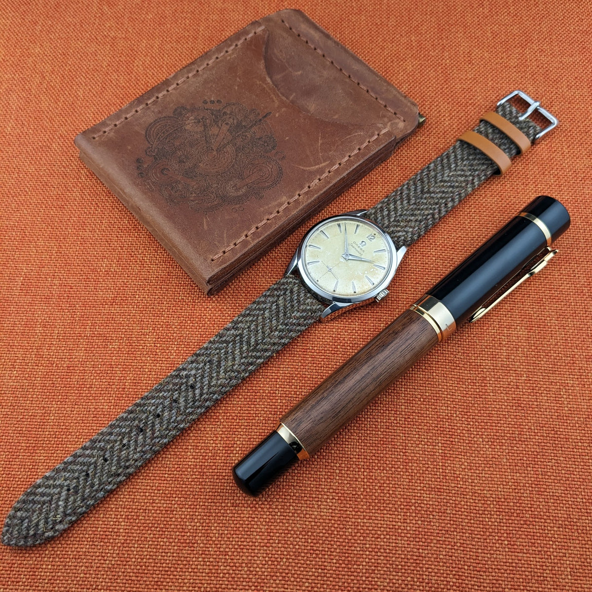 Omega Seamaster reference 29374 with long woven strap arranged beside a brown leather case and black-and-wood pen on orange fabric.