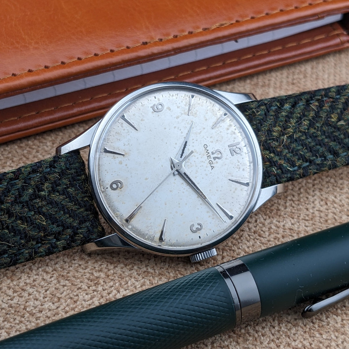 Nahaufnahme des Omega Classic Referenz 2810-2 SC Zifferblatts und grünen Armbands, mit einem braunen Notizbuch und dunklem Stift in der Nähe.
