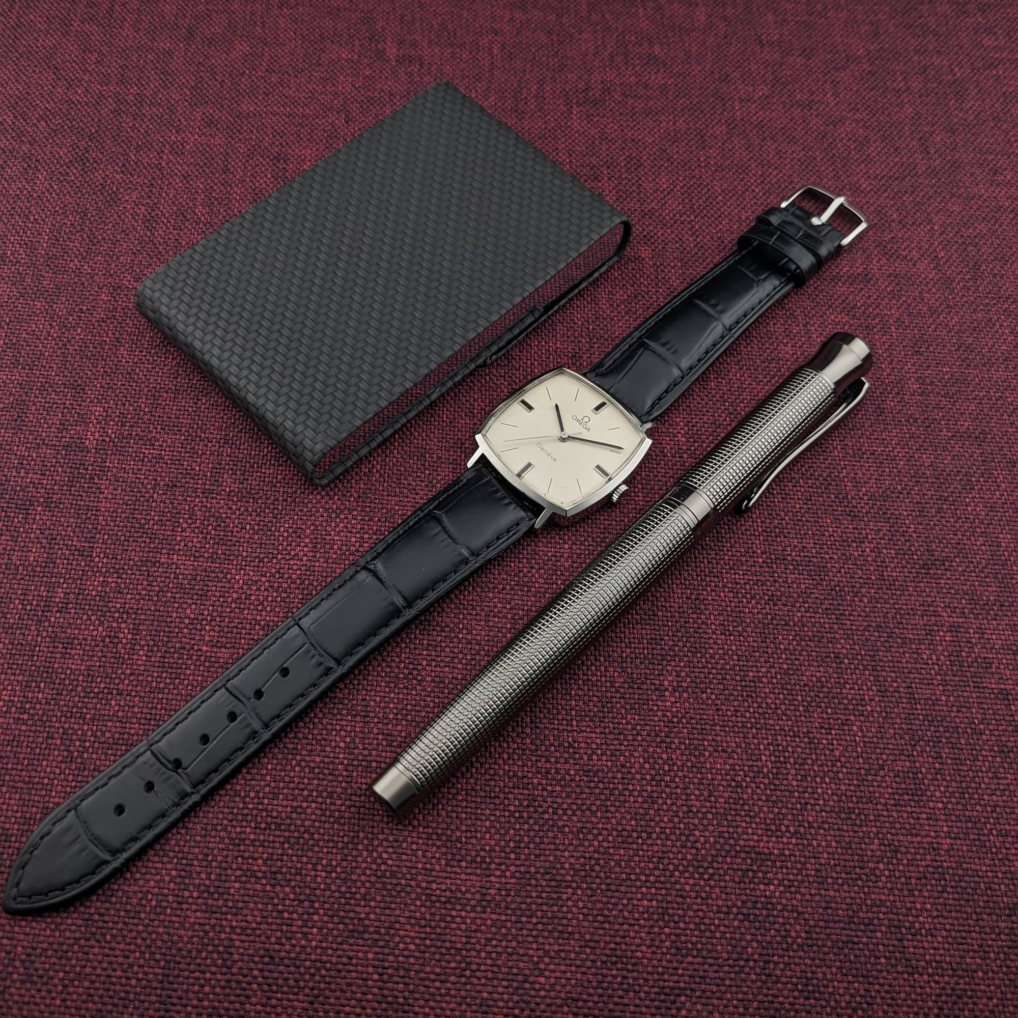 Omega Genève reference 131.022, black leather strap, dark case, and silver pen arranged on burgundy fabric in an overhead composition.