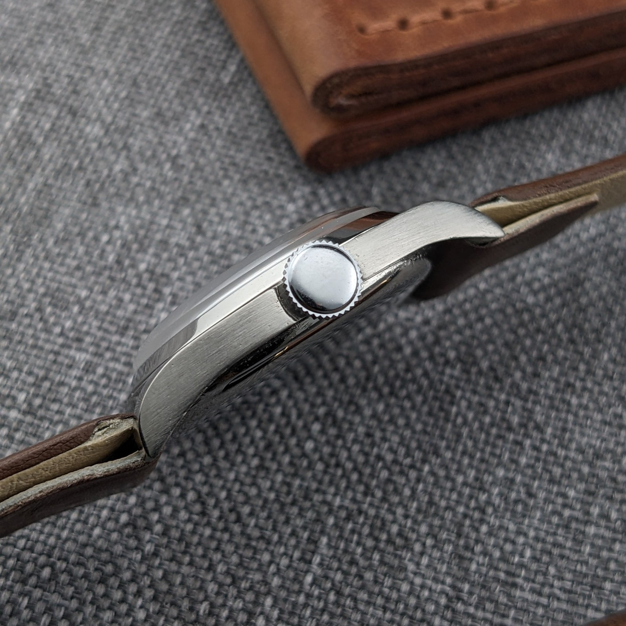 Side profile of an Omega watch showing its rounded case, winding crown, lugs, and brown leather strap against gray fabric.