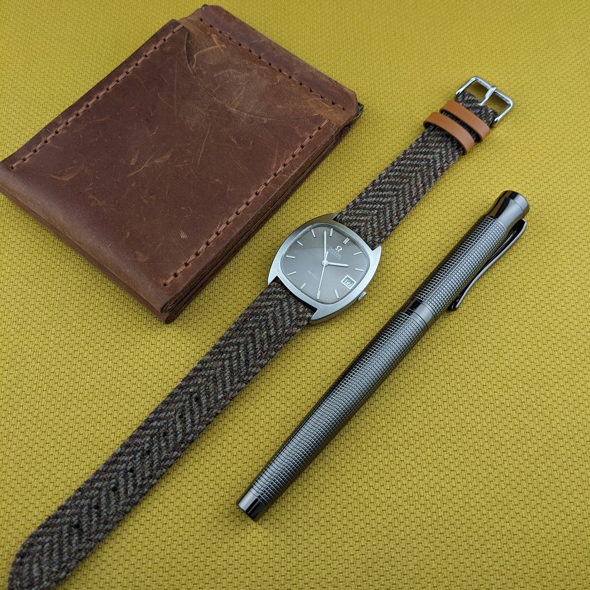 Overview of the Omega Dr Ville Automatic ref. 162.045 with its woven strap, leather wallet, and metal pen arranged on yellow fabric.
