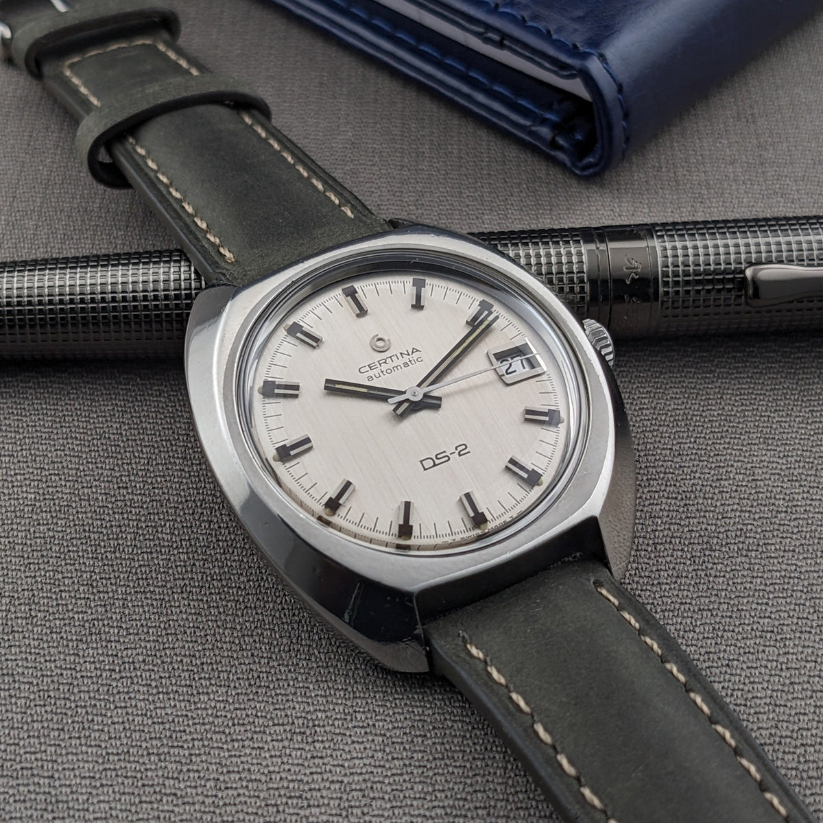 Certina DS-2 Automatic reference 5801 300 positioned on gray fabric beside a textured metal pen and blue wallet, with its full strap visible.