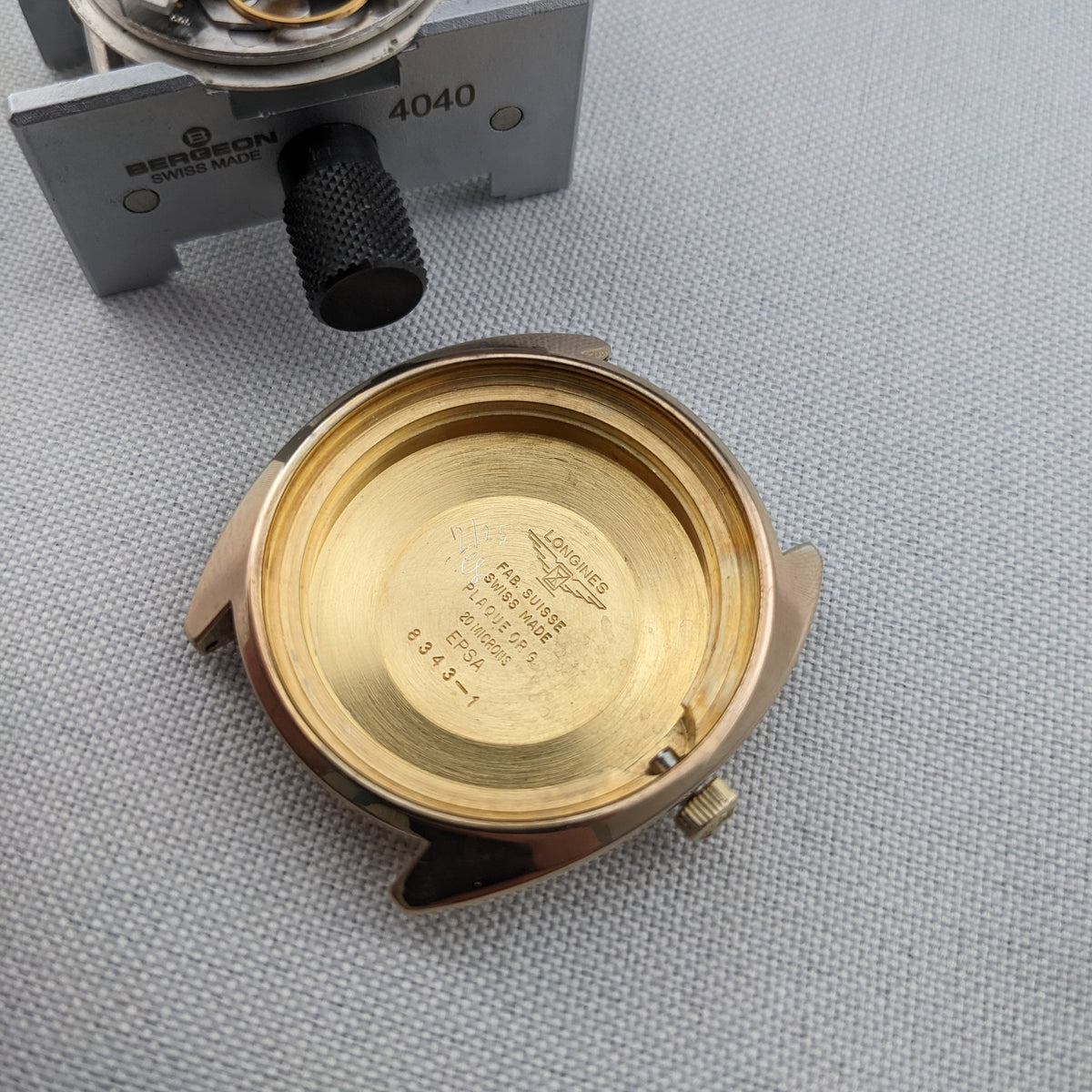 Offenes Longines Admiral 8343-1 Gehäuse von hinten gezeigt, zeigt das goldfarbene Innere und gravierte zentrale Markierungen neben einem Uhrmacherwerkzeug.