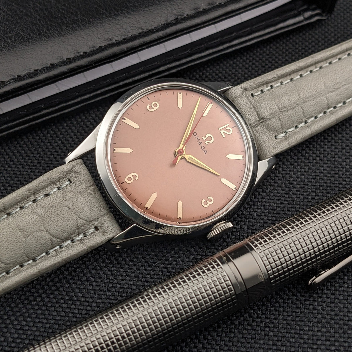 Omega Classic 2760-6 SC neben strukturiertem Metallstift und schwarzem Portemonnaie, mit rosafarbenem Zifferblatt und grauem Armband im Bild.
