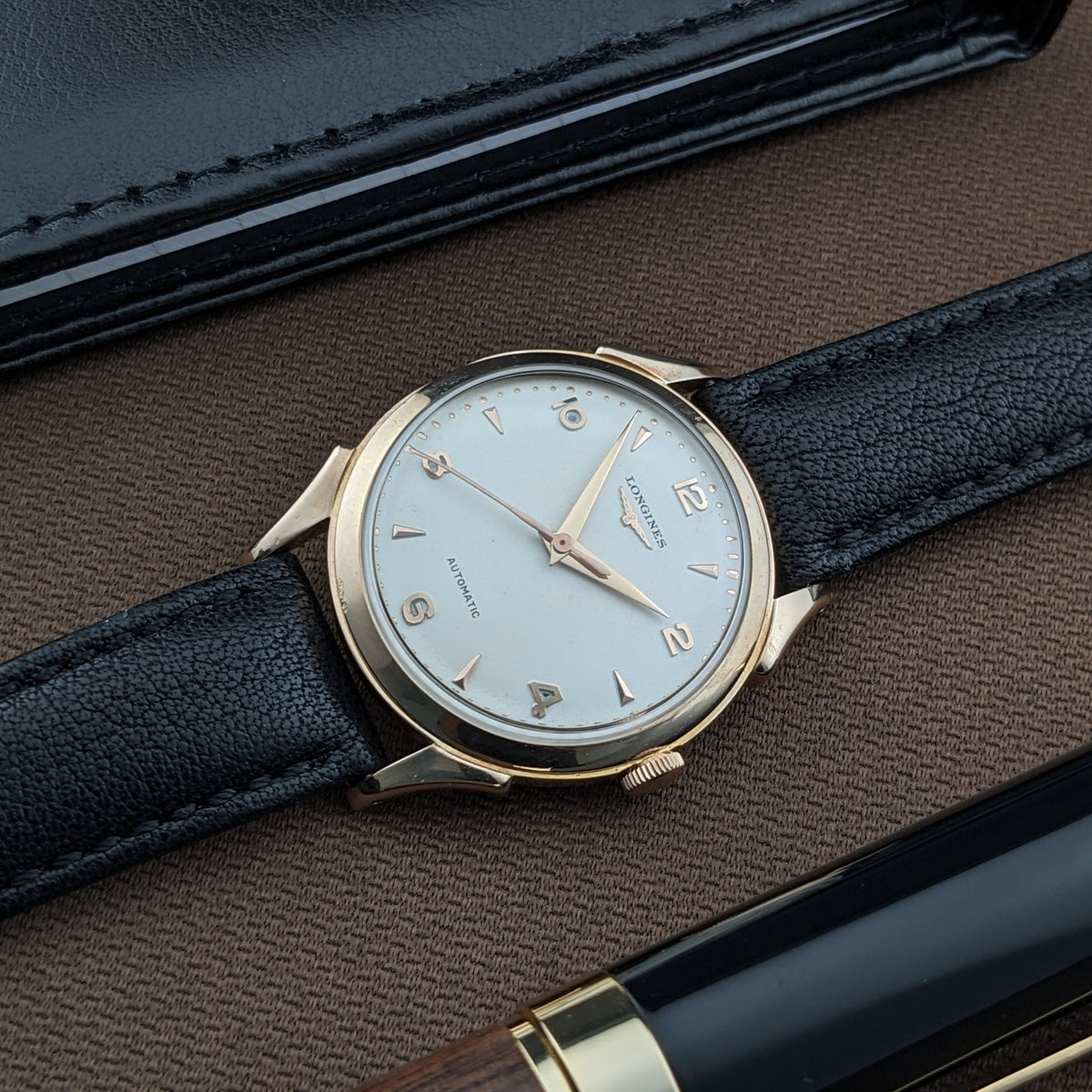 Longines Automatik Referenz 6058 auf braunem Stoff neben einem schwarzen Portemonnaie und Stift, mit sichtbarem hellem Zifferblatt und Armband.