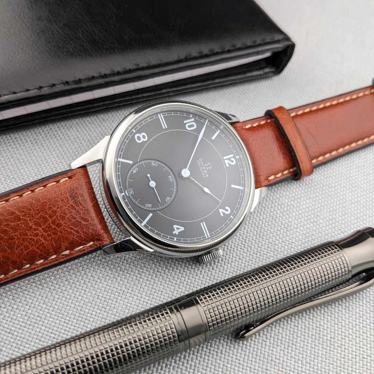 Omega-Armbanduhr liegt horizontal mit dunklem Zifferblatt und braunem genähtem Armband, über einem silbernen Stift und unter einem schwarzen Etui.