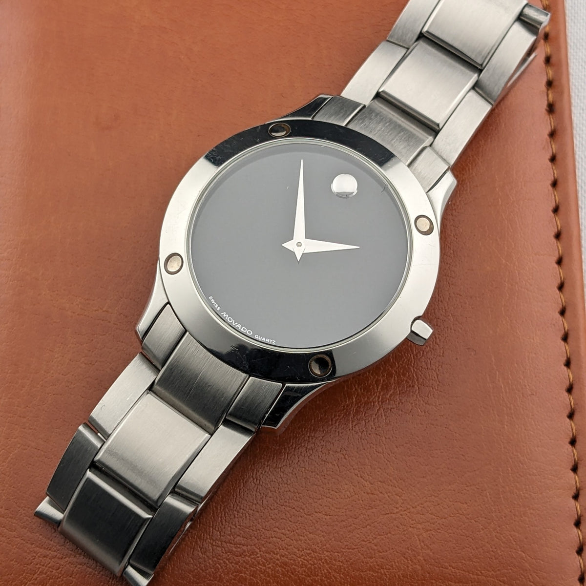 Montre Movado reposant sur un étui en cuir brun, avec cadran noir, aiguilles argentées, repère circulaire, attaches de lunette et bracelet.