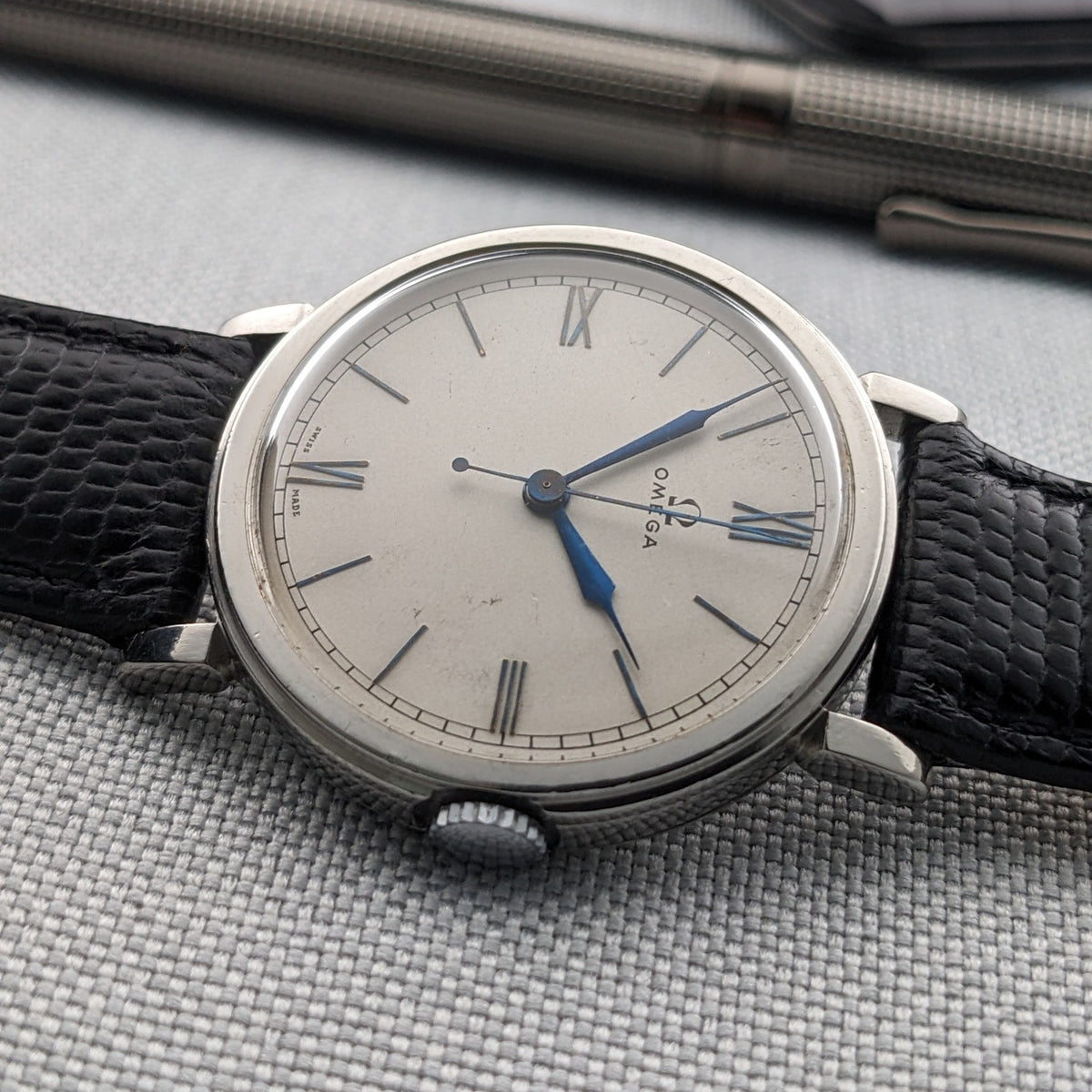 Omega watch photographed sideways with its light dial, blue hands, Roman numerals and black strap on gray fabric.