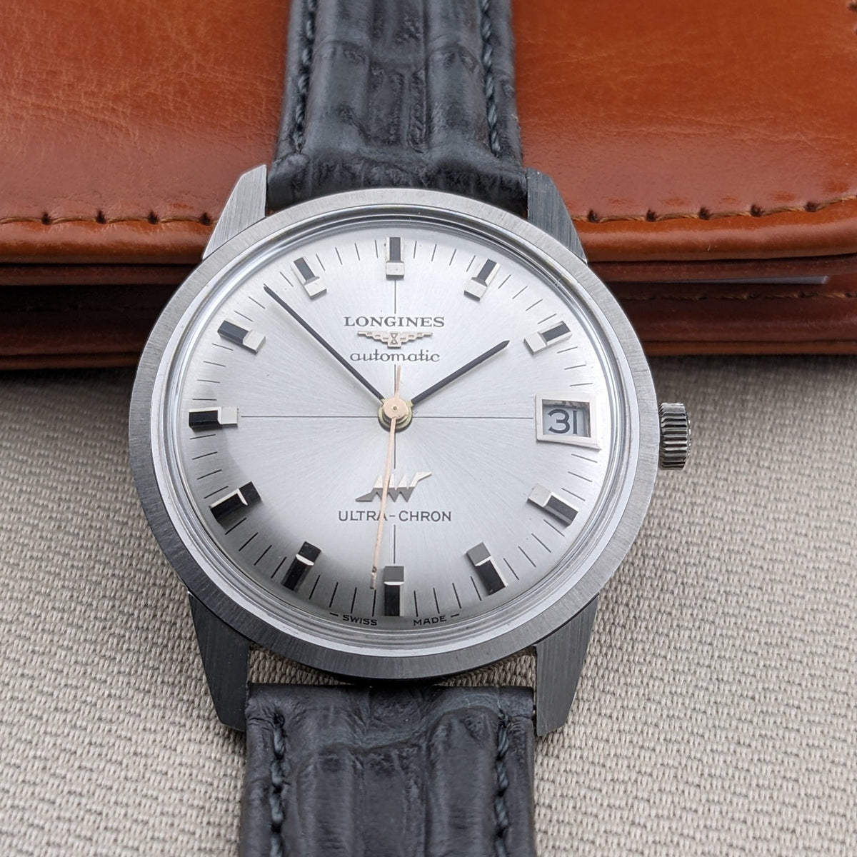 Straight-on view of the Longines Ultra-Chron 7827 with silver dial, applied markers, date window, black strap, and a brown leather case behind.