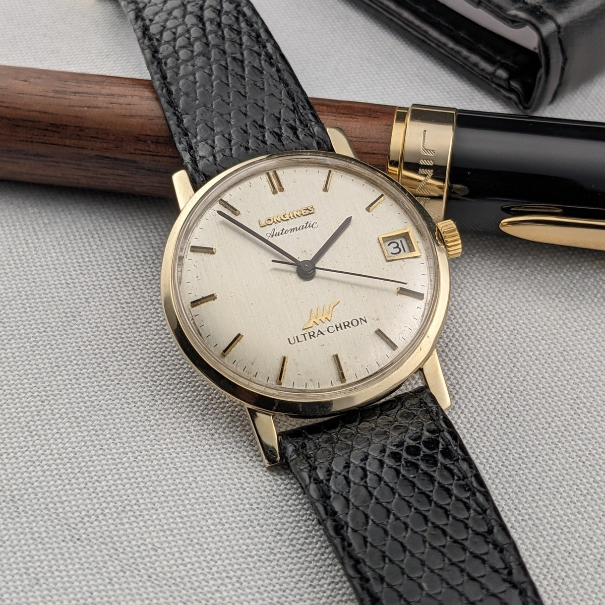 Longines Ultra-Chron reference 69-556AC shown diagonally beside a black-and-wood pen, with its dial and textured strap prominent.