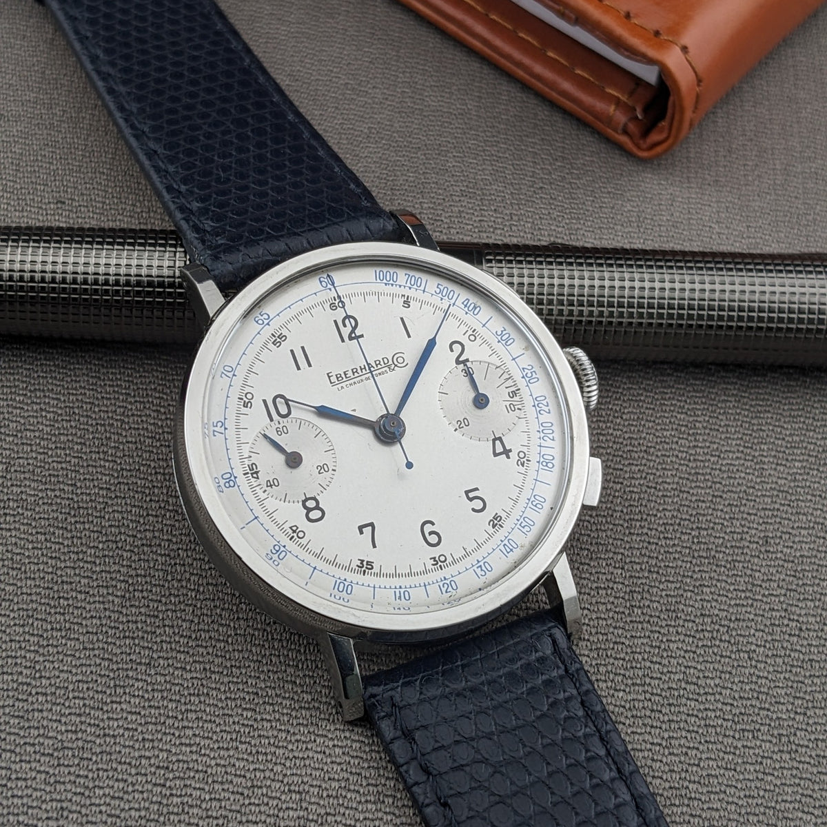 Eberhard & Co.. Pre-Extra Fort lies diagonally across gray fabric with its cream chronograph dial, blue strap, wallet and pen visible.
