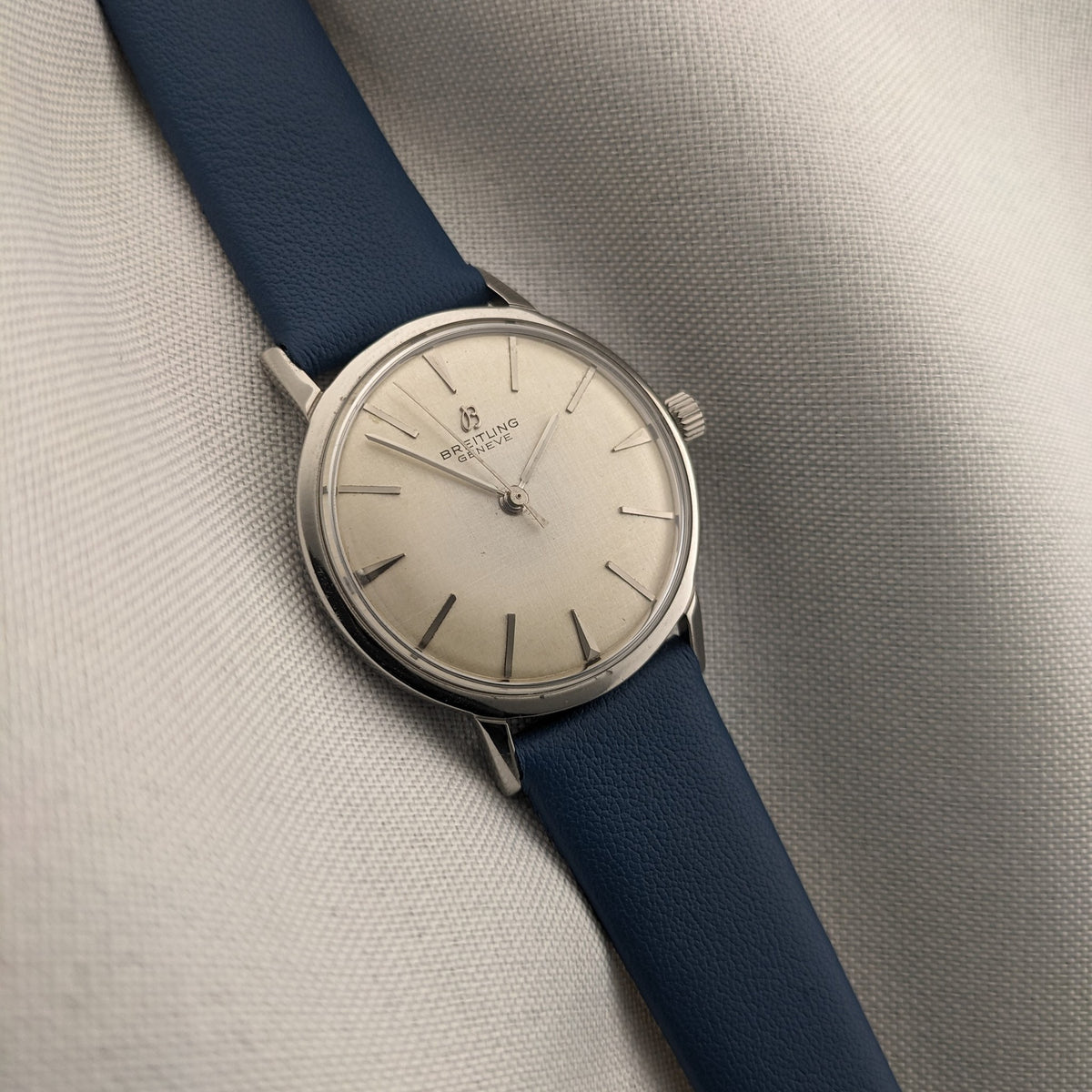 Breitling Automatic watch lies diagonally on a light textured surface, showing its cream dial, blue strap and polished round case.