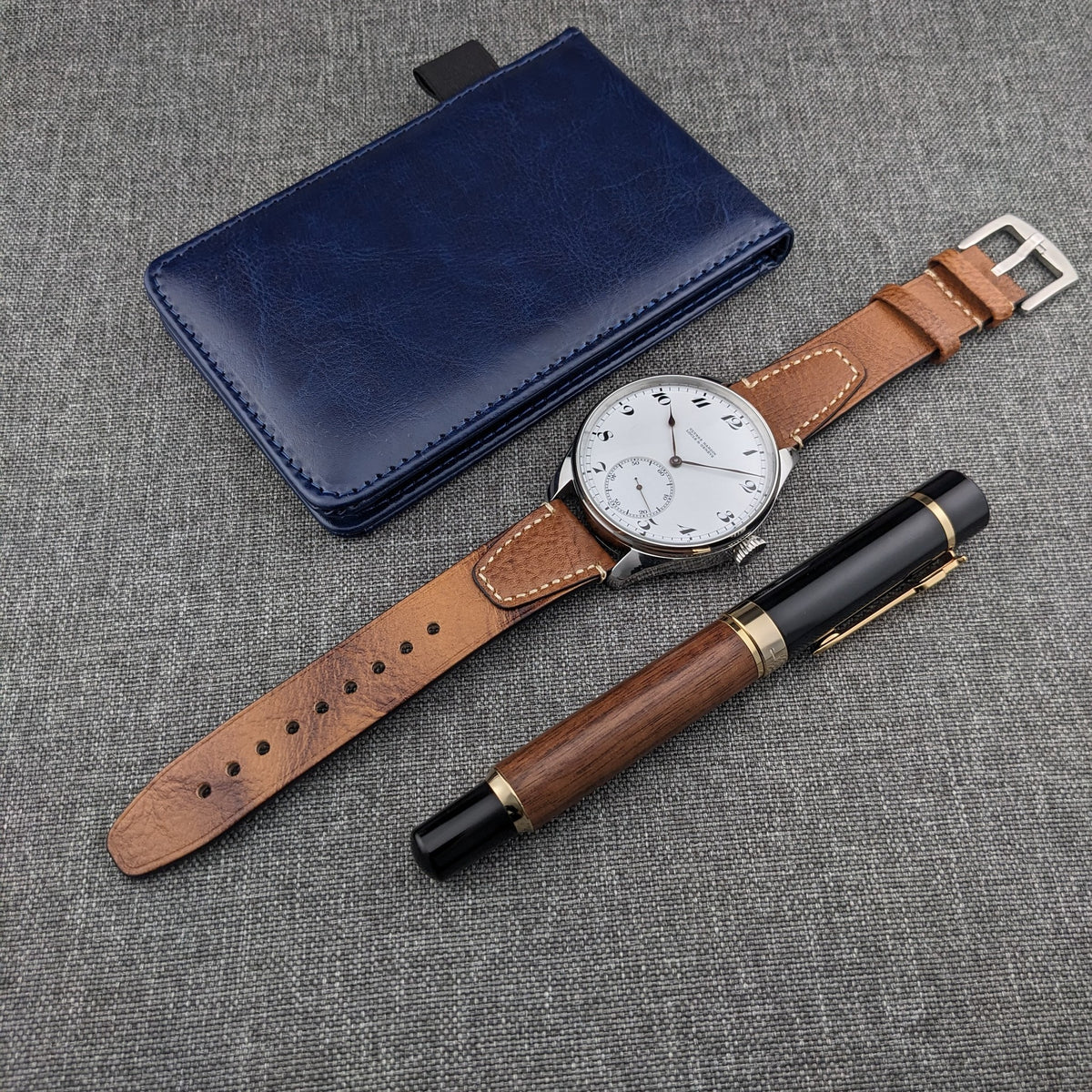 Ulysse Nardin कलाई घड़ी, विस्तारित भूरा चमड़े का पट्टा, नीला चमड़े का केस और दो लेखन उपकरण ग्रे कपड़े पर व्यवस्थित।