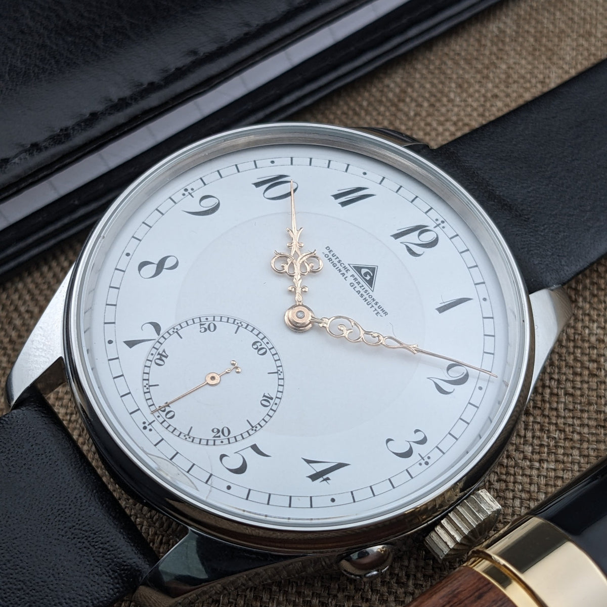 Schrägansicht der Deutsche Präzisionsuhr „Original Glashütte” mit weißem Zifferblatt, verzierten Zeigern, arabischen Ziffern