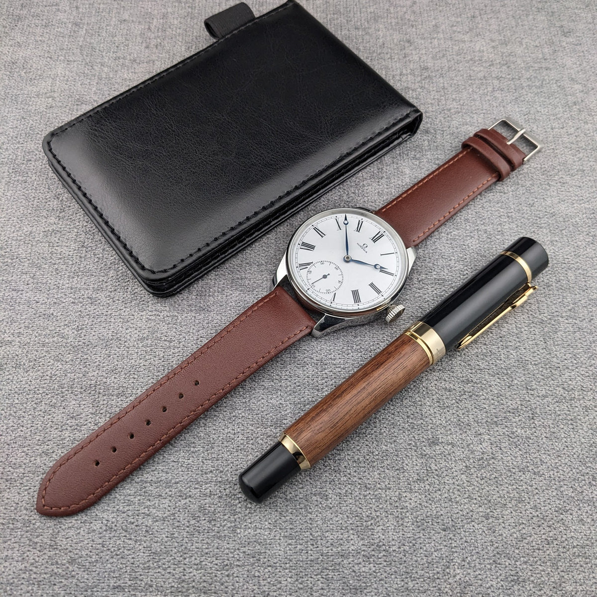 Orologio Omega, custodia in pelle nera e penna in legno e nero disposti insieme su tessuto grigio, con cinturino marrone completamente visibile.