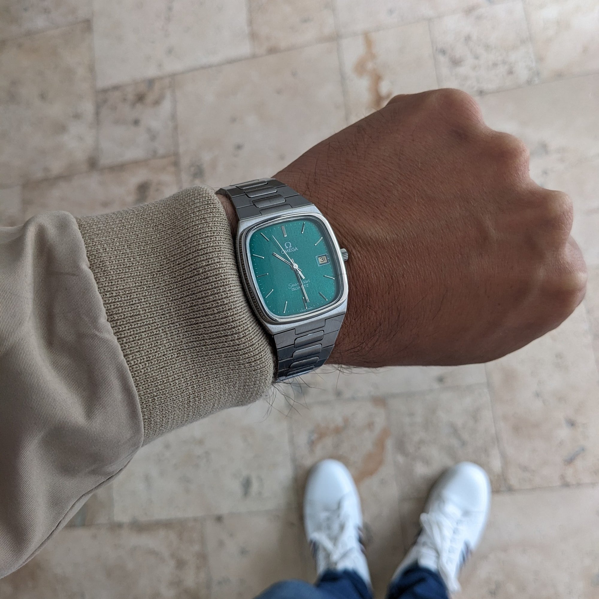 Wrist-level view of the Omega Seamaster reference 196.0166 with teal dial and metal bracelet against a pale tiled floor.