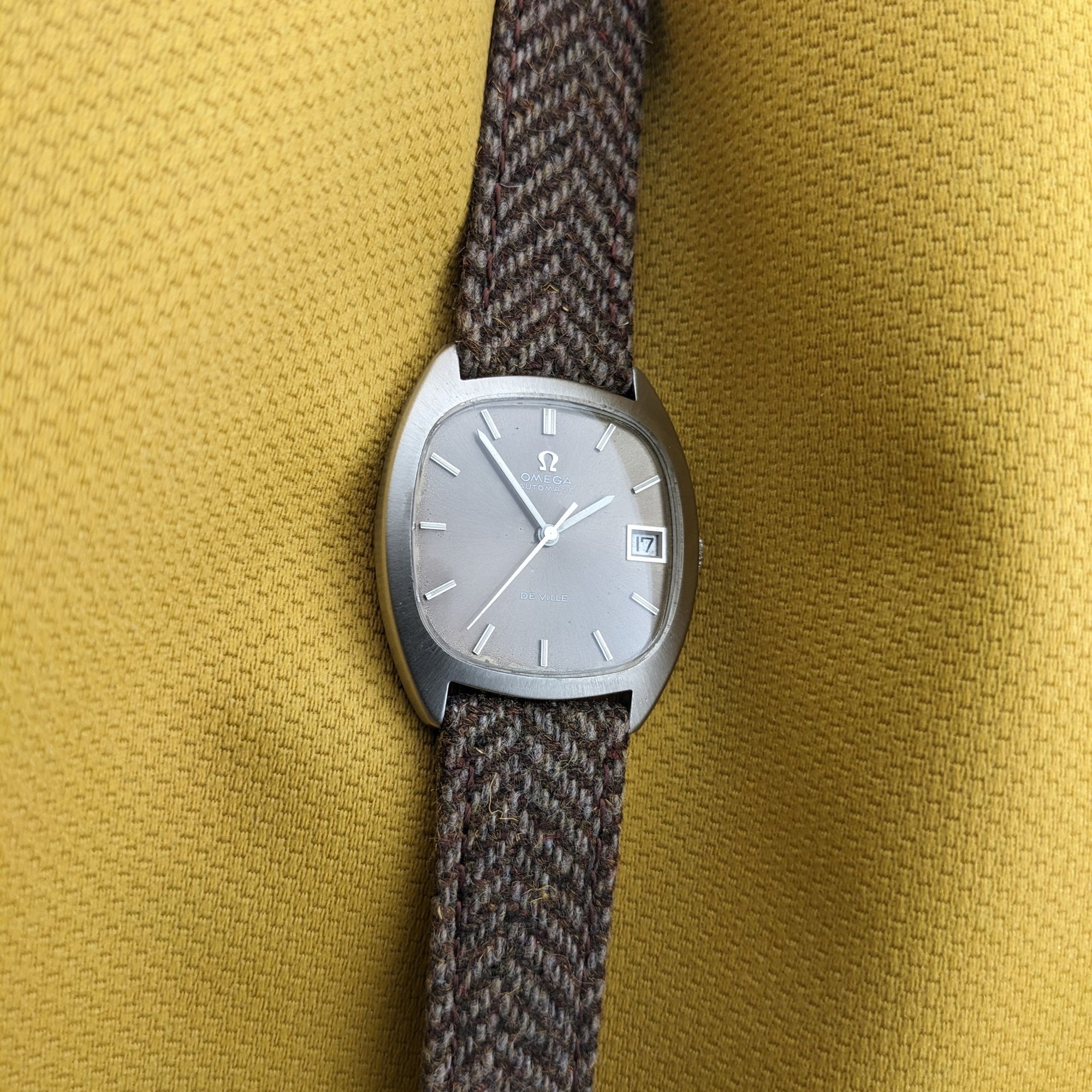 Straight-on view of the Omega Dr Ville Automatic ref. 162.045 centered on yellow fabric, showing the silver dial, date window, and woven strap.