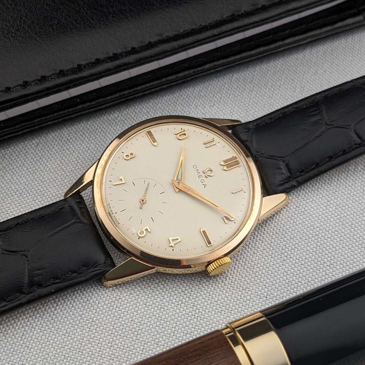 Angled front view of the Omega Classic reference 14391-1 on fabric, with the light dial, gold-tone case and black strap beside a pen.