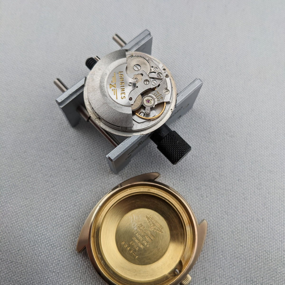 Longines Admiral Referenz 8343-1 Gehäuse und freigelegtes mechanisches Werk separat auf einer strukturierten Oberfläche mit einem Uhrenhalter gezeigt.