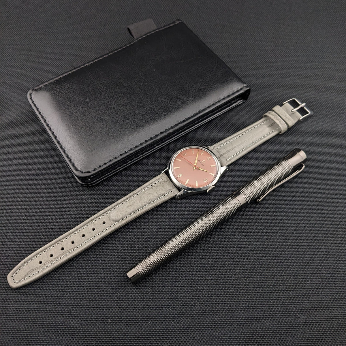 Omega Classic Referenz 2760-6 SC flach liegend mit ausgezogenem grauem Armband, begleitet von schwarzem Portemonnaie und strukturiertem Metallstift.