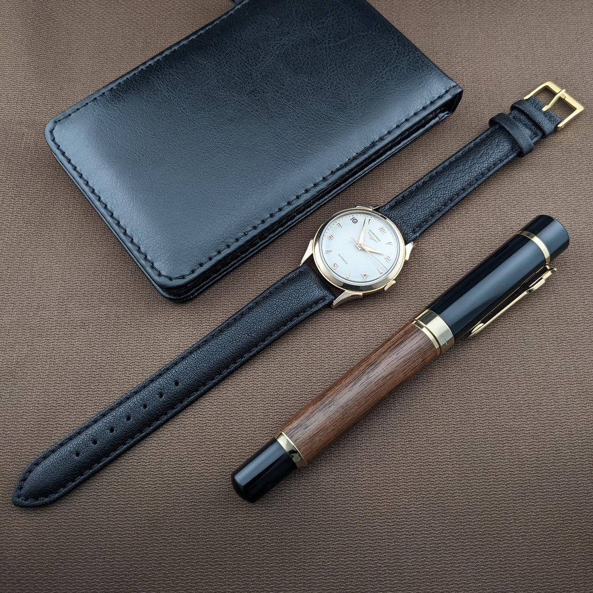 Longines Automatik Referenz 6058 mit schwarzem Lederportemonnaie und Holz-Schwarz-Stift auf braunem Stoff.