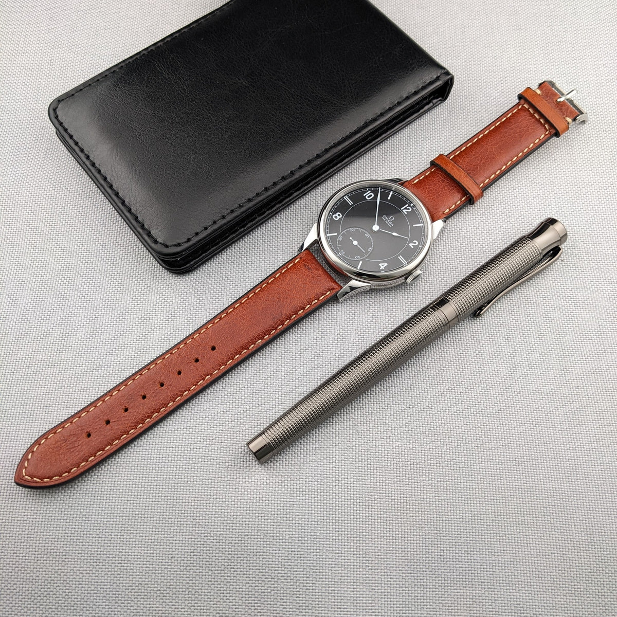 Omega-Armbanduhr, verlängertes braunes Lederarmband, schwarzes Etui und silberner Stift zusammen auf einer hell strukturierten Oberfläche arrangiert.