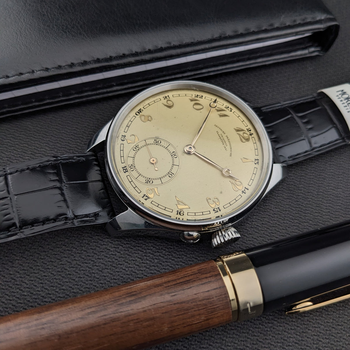 Schrägansicht der Deutsche Präzisions-Uhrenfabrik Glashütte Uhr neben einer schwarzen Geldbörse und einem Holz-und-schwarzen Stift, mit sichtbarem Zifferblatt